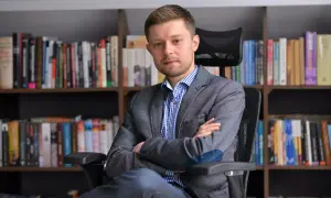 Mężczyzna w marynarce siedzi na krześle przed regałami pełnymi książek. Zastanawiasz się, ile książek ma Remigiusz Mróz?