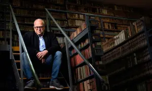 Mężczyzna w okularach siedzi na schodach w bibliotece pełnej starych ksiąg, jakby szukał inspiracji do romantycznych wierszy na dobranoc.