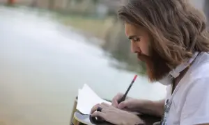 Mężczyzna z brodą i długimi włosami pisze wiersz dla ukochanej kobiety, siedząc przy gitarze nad wodą.