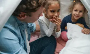 Tata opowiada dziewczynkom śmieszne rymy krótkie w namiocie z kocy. Jedna z córek chichocze, zakrywając usta.