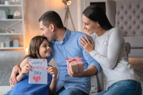Córka wręcza tacie kartkę z napisem "Happy Father's Day" i prezent. Mama obejmuje męża. Piękny wiersz dla męża czeka na odczytanie.