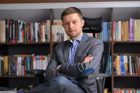 Mężczyzna w marynarce siedzi na krześle przed regałami pełnymi książek. Zastanawiasz się, ile książek ma Remigiusz Mróz?