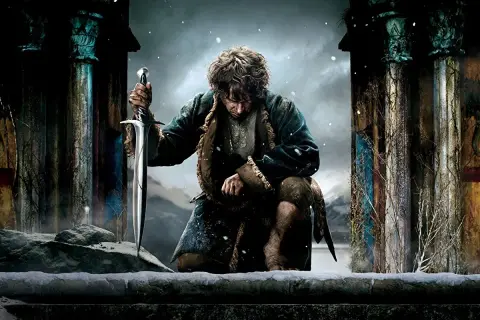 Bilbo Baggins, jak wygląda hobbit, klęczy w śniegu, trzymając miecz. W tle ruiny i zimowy krajobraz.