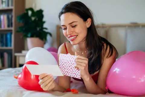 Uśmiechnięta kobieta czyta kartkę z serduszkami, otoczona balonami. Może to być inspiracja na wiersze dla ukochanego mężczyzny.