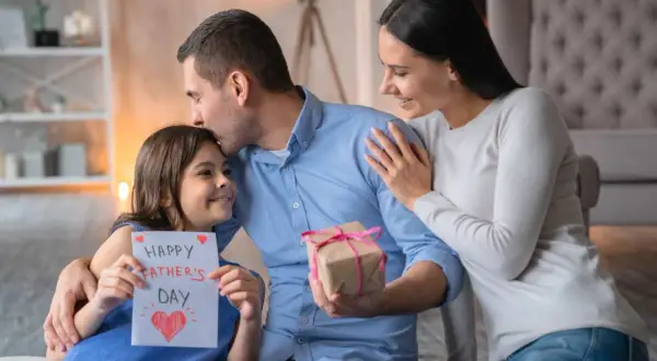 Córka wręcza tacie kartkę z napisem "Happy Father's Day" i prezent. Mama obejmuje męża. Piękny wiersz dla męża czeka na odczytanie.