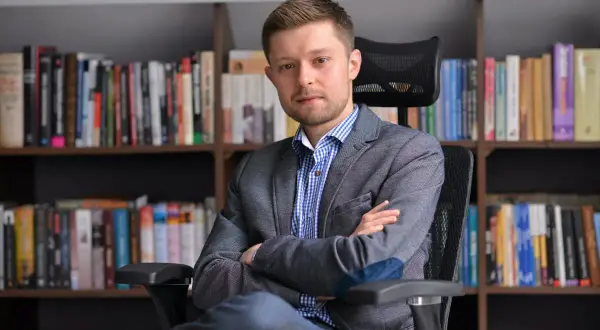 Mężczyzna w marynarce siedzi na krześle przed regałami pełnymi książek. Zastanawiasz się, ile książek ma Remigiusz Mróz?