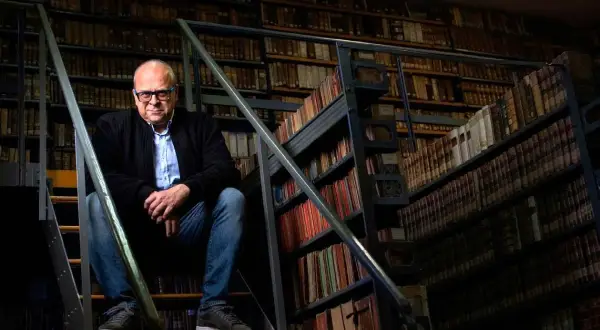Mężczyzna w okularach siedzi na schodach w bibliotece pełnej starych ksiąg, jakby szukał inspiracji do romantycznych wierszy na dobranoc.