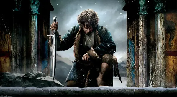 Bilbo Baggins, jak wygląda hobbit, klęczy w śniegu, trzymając miecz. W tle ruiny i zimowy krajobraz.