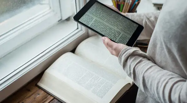 Osoba porównuje tekst na tablecie z otwartą księgą. Czy warto czytać książki? Przykłady z literatury inspirują do poszukiwań.