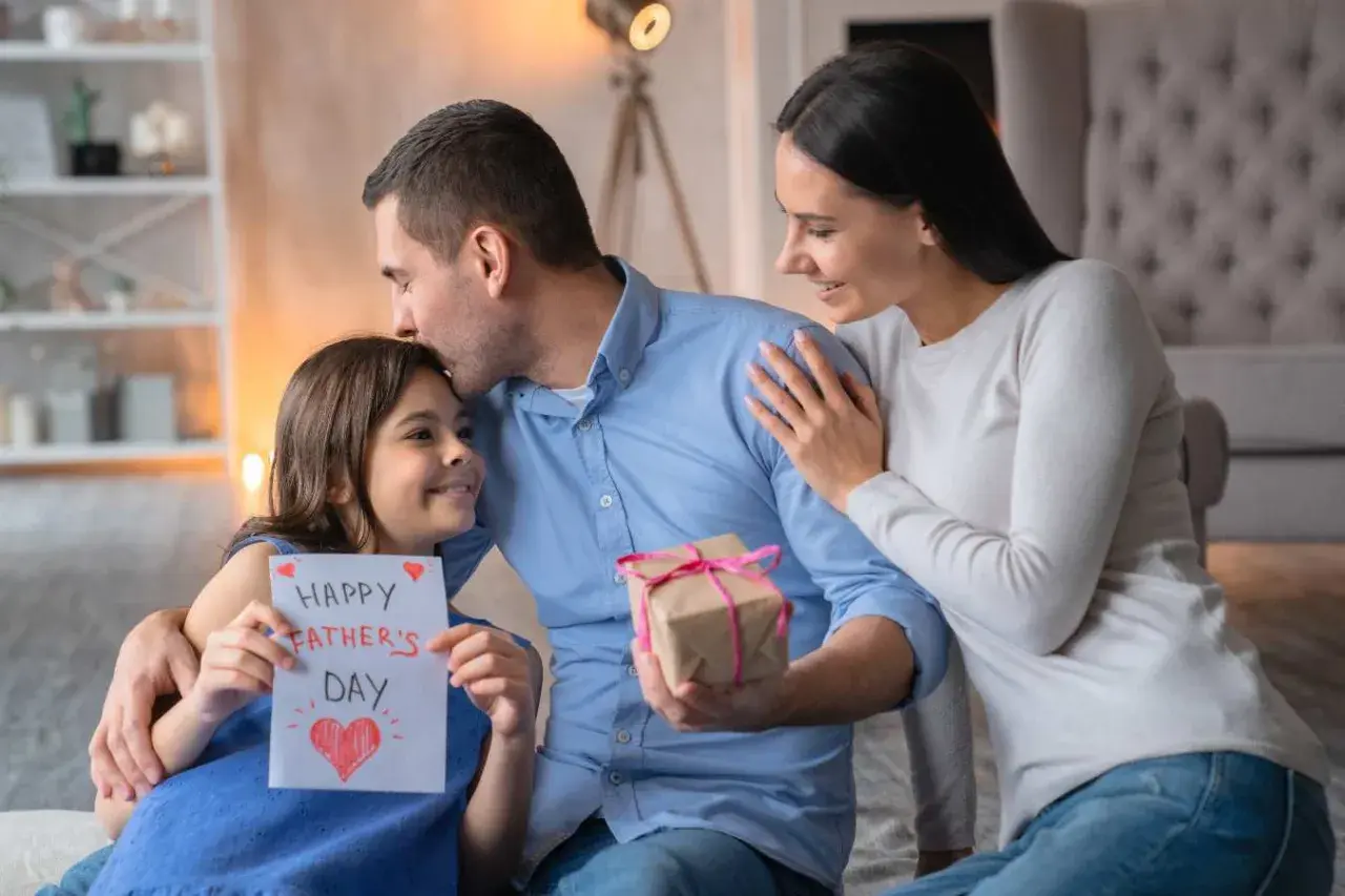 Córka wręcza tacie kartkę z napisem "Happy Father's Day" i prezent. Mama obejmuje męża. Piękny wiersz dla męża czeka na odczytanie.