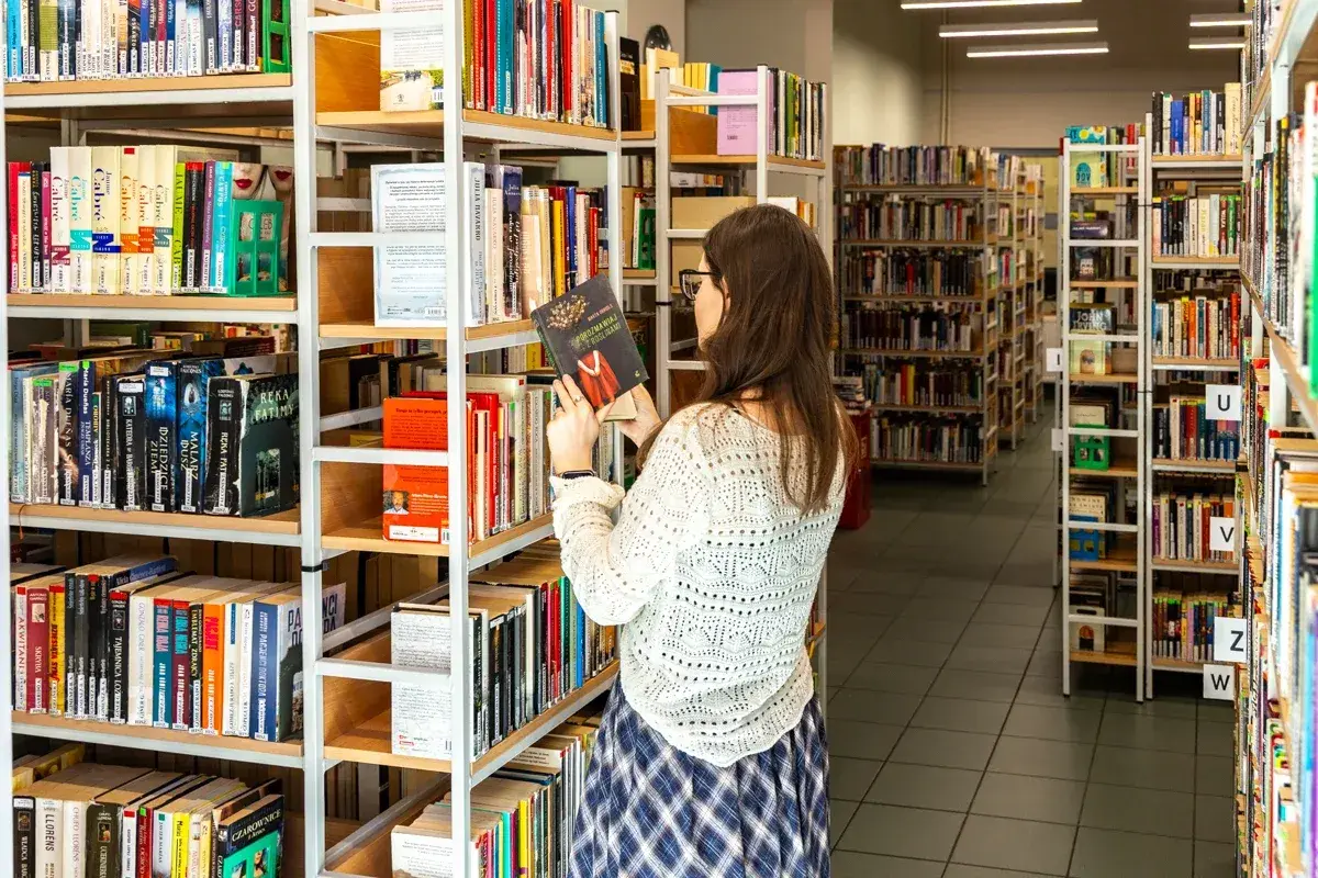 W bibliotece, gdzie **miejscu możecie znaleźć szeroki** wybór książek, młoda kobieta przegląda półki, trzymając w ręku jedną z pozycji.