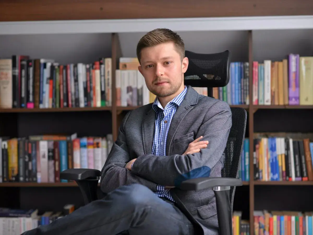 Mężczyzna w marynarce siedzi na krześle przed regałami pełnymi książek. Zastanawiasz się, ile książek ma Remigiusz Mróz?