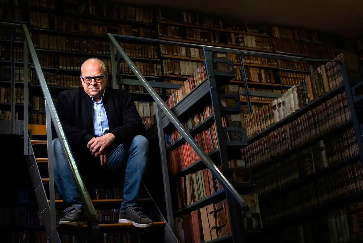 Mężczyzna w okularach siedzi na schodach w bibliotece pełnej starych ksiąg, jakby szukał inspiracji do romantycznych wierszy na dobranoc.