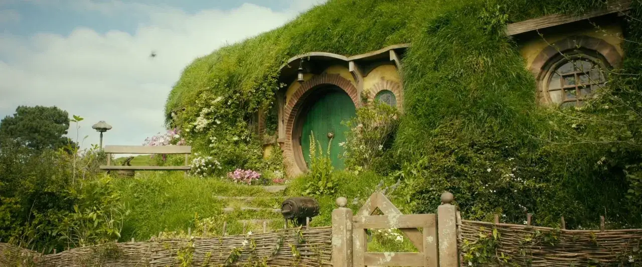 Zielone drzwi i okrągłe okna w zboczu wzgórza, gdzie mieszkał Bilbo Baggins. Wokół bujna zieleń i kwiaty.
