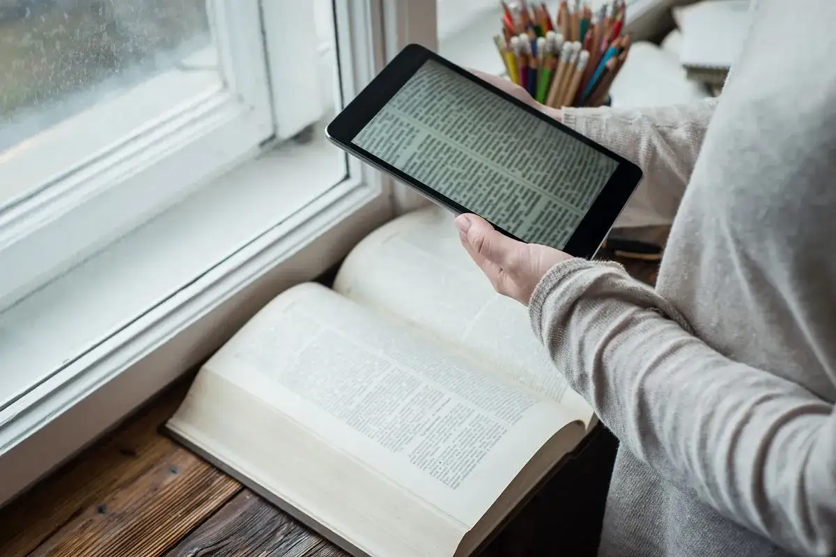 Osoba porównuje tekst na tablecie z otwartą, obszerną księgą. Czy warto czytać książki? Przykłady z literatury mogą pomóc w tej decyzji.