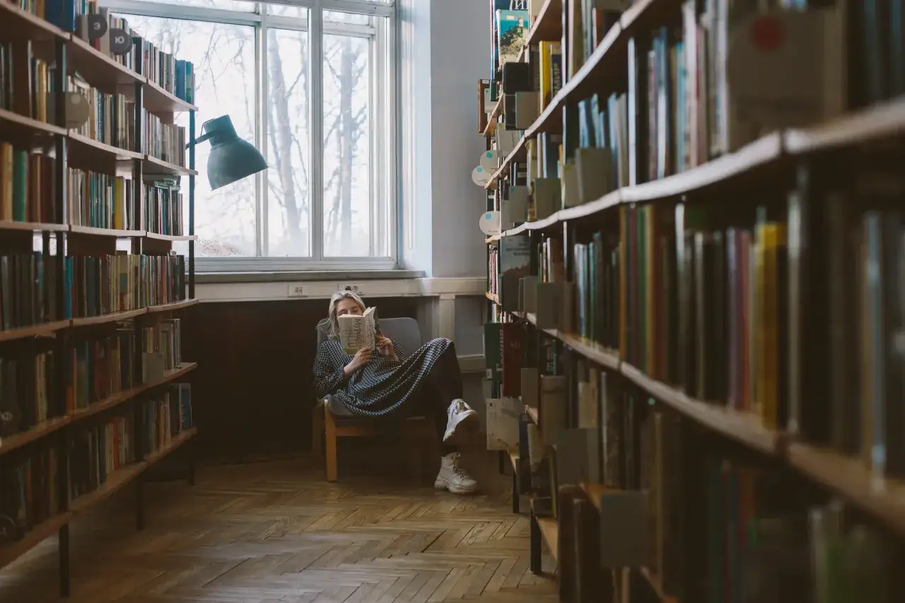 W bibliotece, gdzie można znaleźć szeroki wybór książek, młoda kobieta siedzi w fotelu przy oknie i czyta.