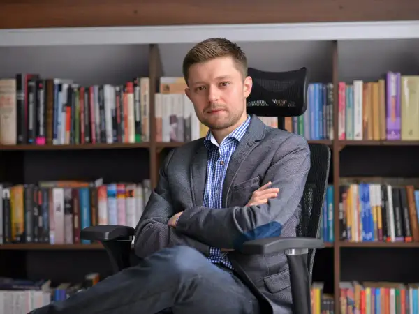 Mężczyzna w marynarce siedzi na krześle przed regałami pełnymi książek. Zastanawiasz się, ile książek ma Remigiusz Mróz?